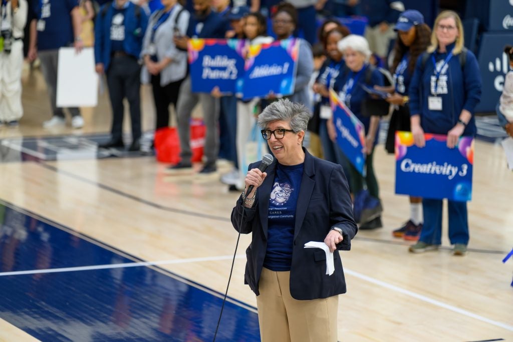 Woman in blue blazer with glasses and gray hair speaks with a mic in a gym