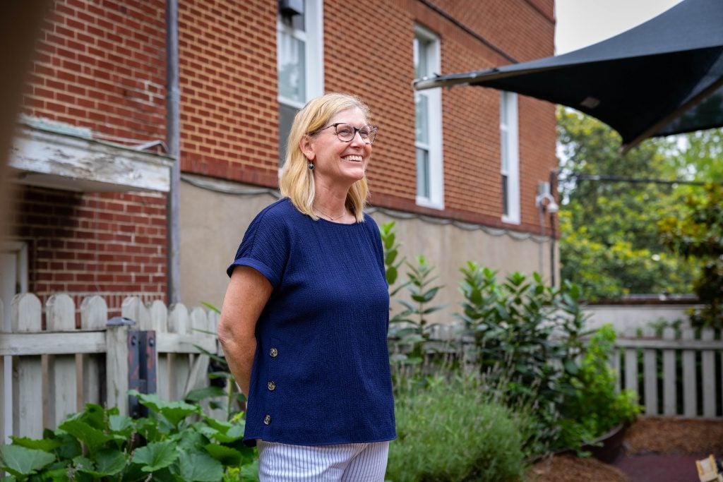 A woman in a navy shirt smiles in front of a brick building and a garden