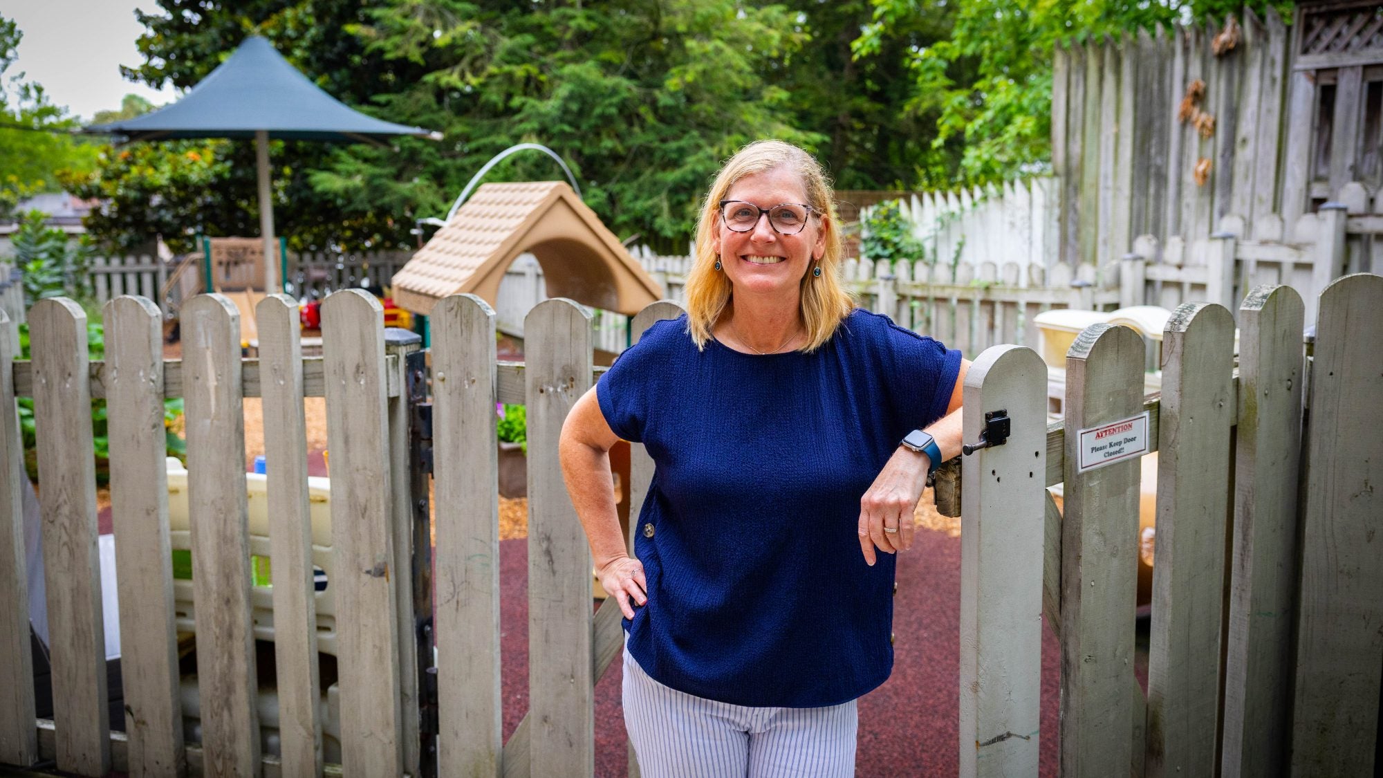 A woman in a navy shirt leans on a fence with a playground behind her