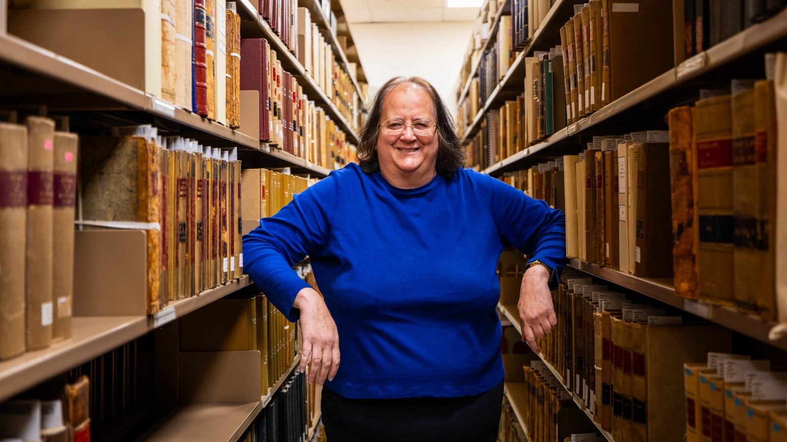 A woman in a bright blue shirt poses in between shelves of archival records