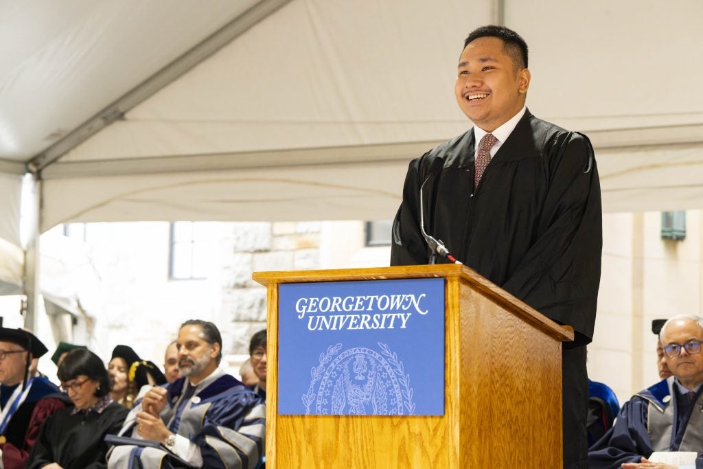 A young Asian man in academic gown smiles at the podium
