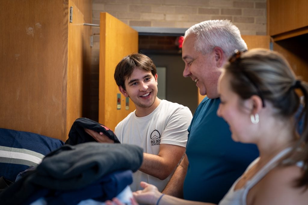 A young man setting up his bed in his dorm with his father and sister
