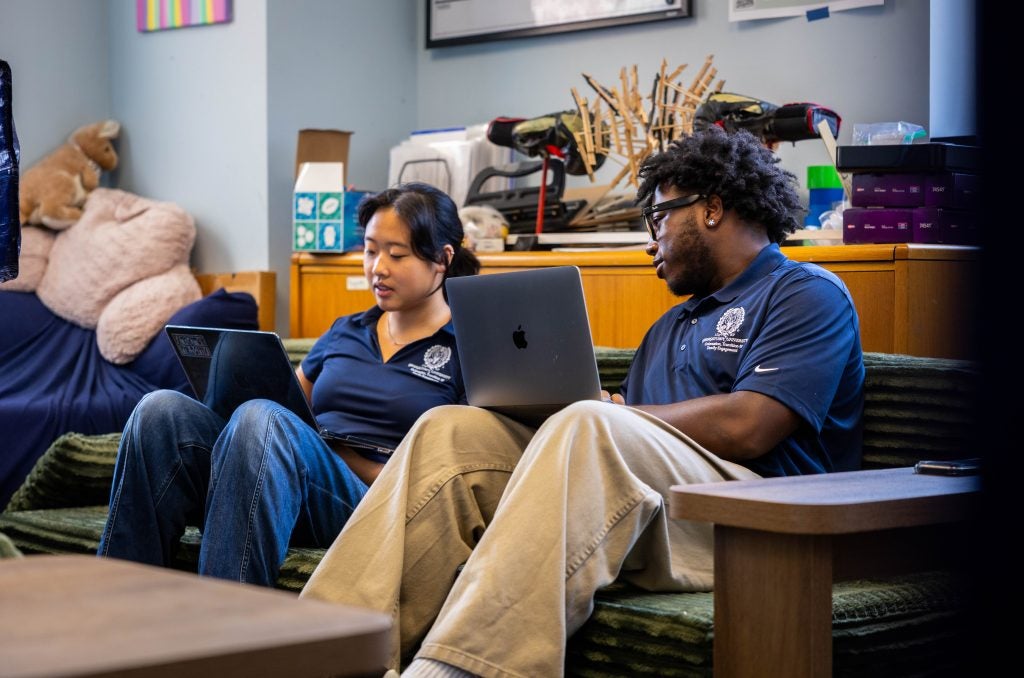 Two students in blue polo shirts chat while sitting with their laptops on their laps