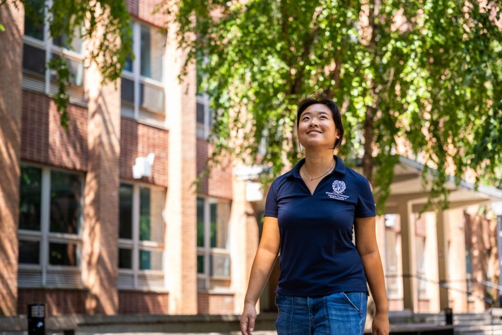 A female student in a blue polo smiles while walking under a tree on a sunny day