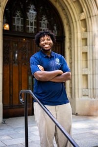 A male student in a blue polo smiles in front of a wooden doorway