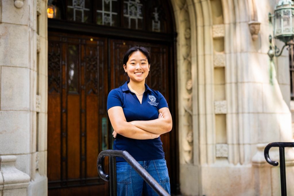 A female student in a blue polo smiles in front of a wooden doorway