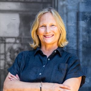 A blonde woman in a navy short-sleeve shirt smiles with her arms crossed