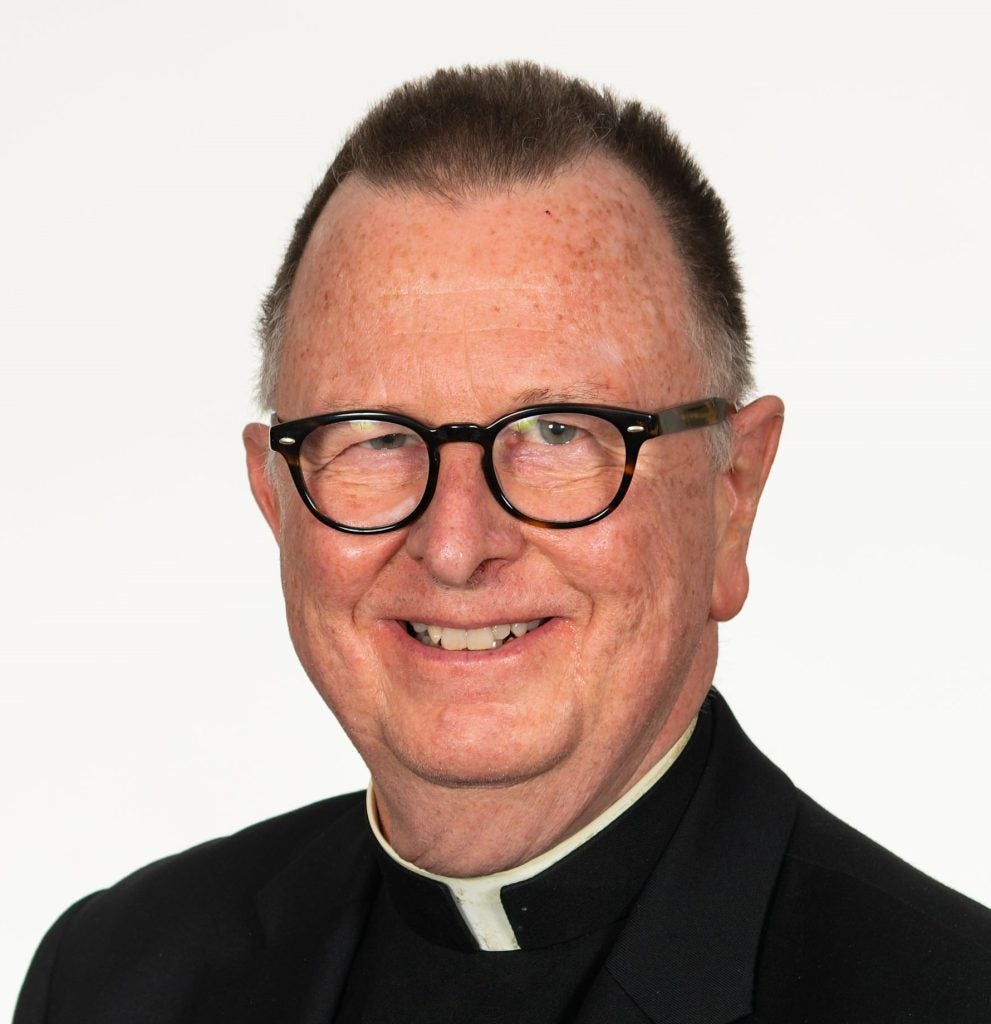 Older Caucasian male with glasses and smiling while wearing a Roman Catholic collar