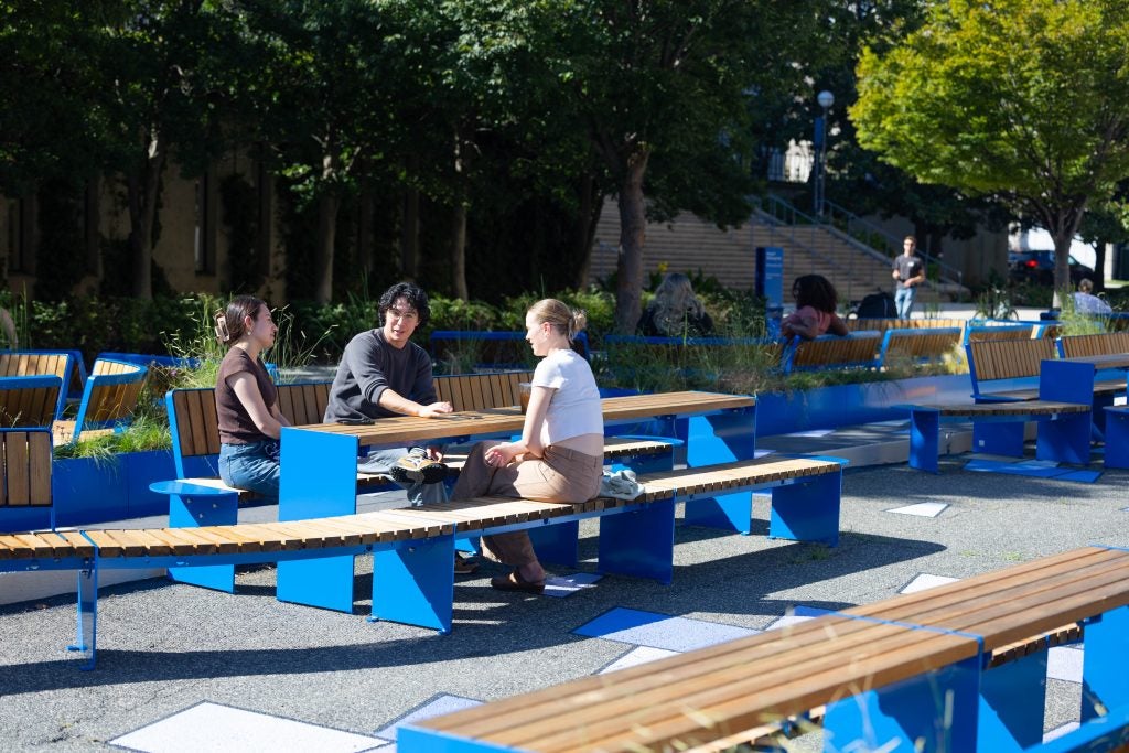 Students sit on wooden, bright blue furniture outdoors