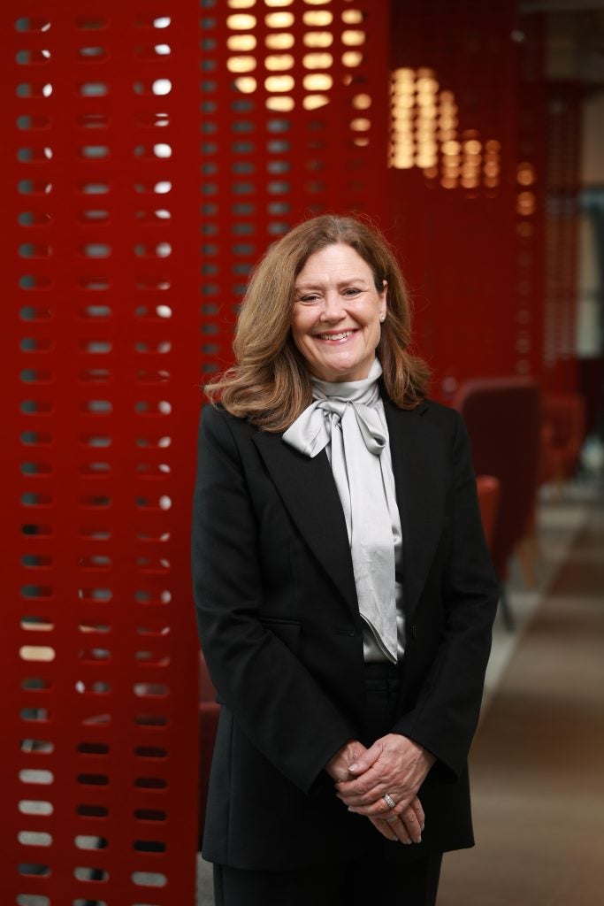 Older Caucasian woman in professional attire and smiling while standing in front of a red background
