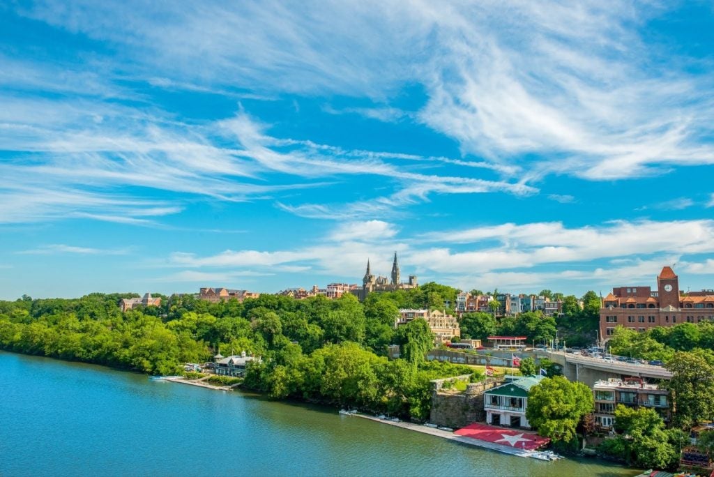 The coastline of the Potomac River with a boathouse on the shore and the university's campus in the background