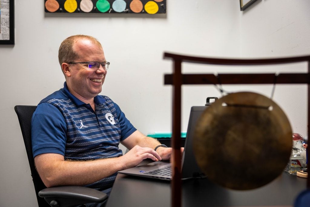 Caucasian man with glasses in blue Georgetown shirt smiling while on his laptop at desk