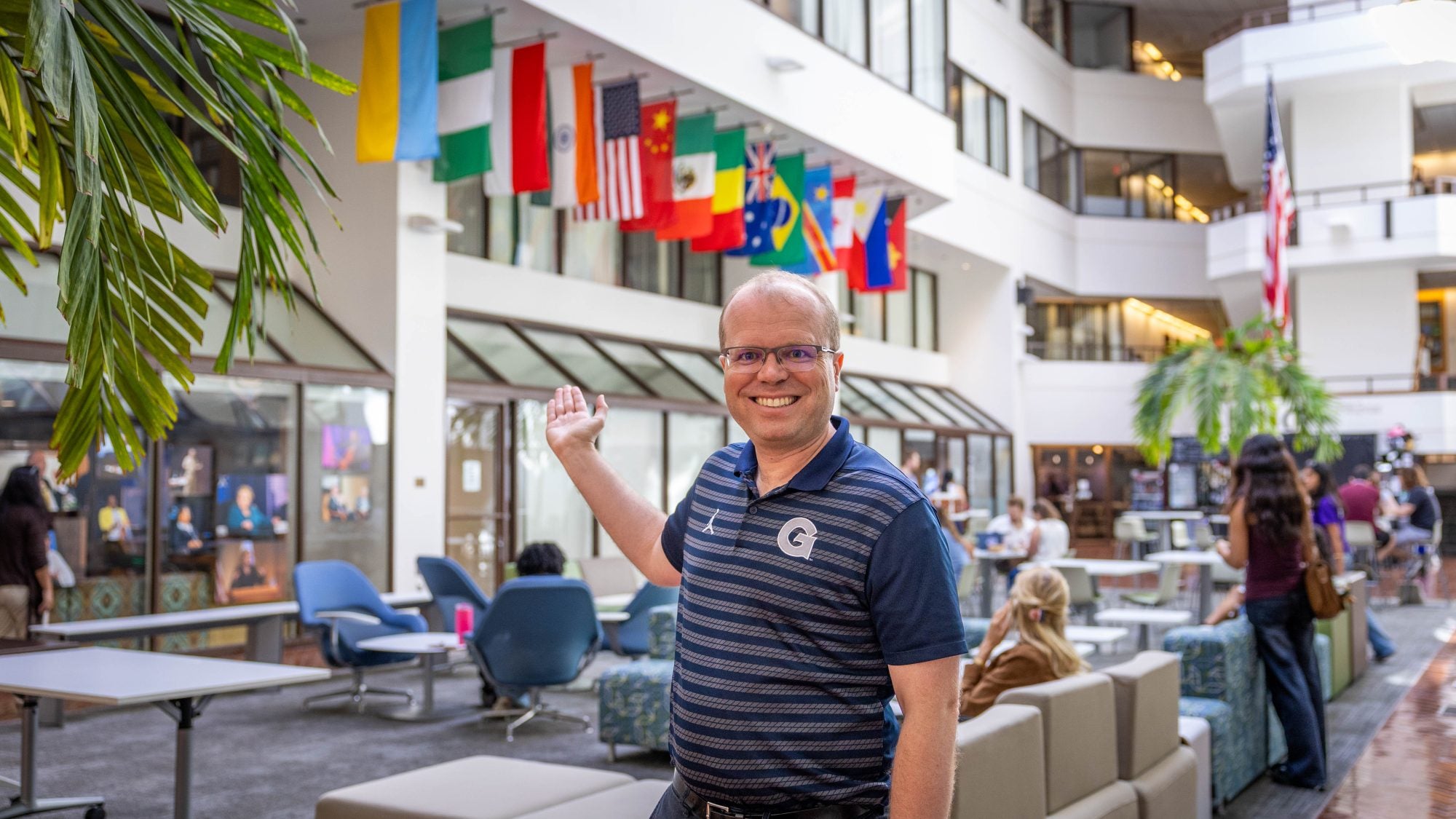 Caucasian man with glasses in blue Georgetown shirt smiling pointing to flags in ICC