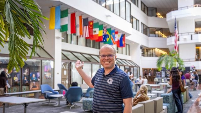 Caucasian man with glasses in blue Georgetown shirt smiling pointing to flags in ICC