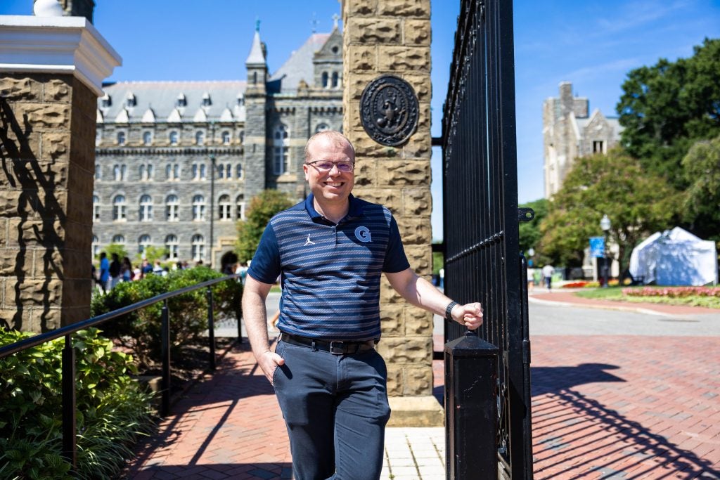 Caucasian man with glasses in blue Georgetown shirt smiling outside of front gates at GU