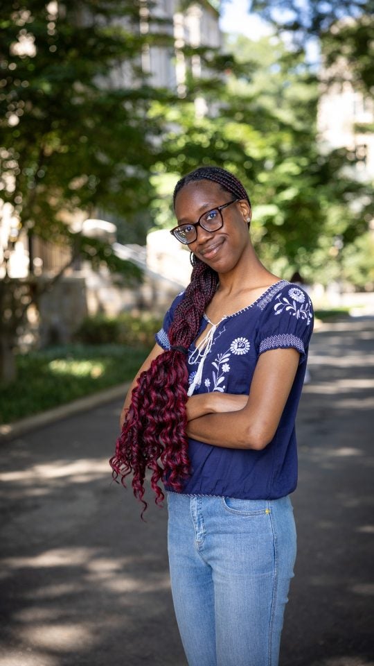 Young Black woman in blue shirt smiling with arms crossed around Copley Lawn
