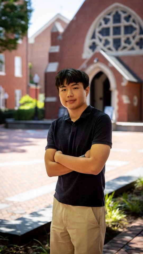 Young Asian man in dark blue shirt with arms crossed and smiling, Dahlgren Chapel in background