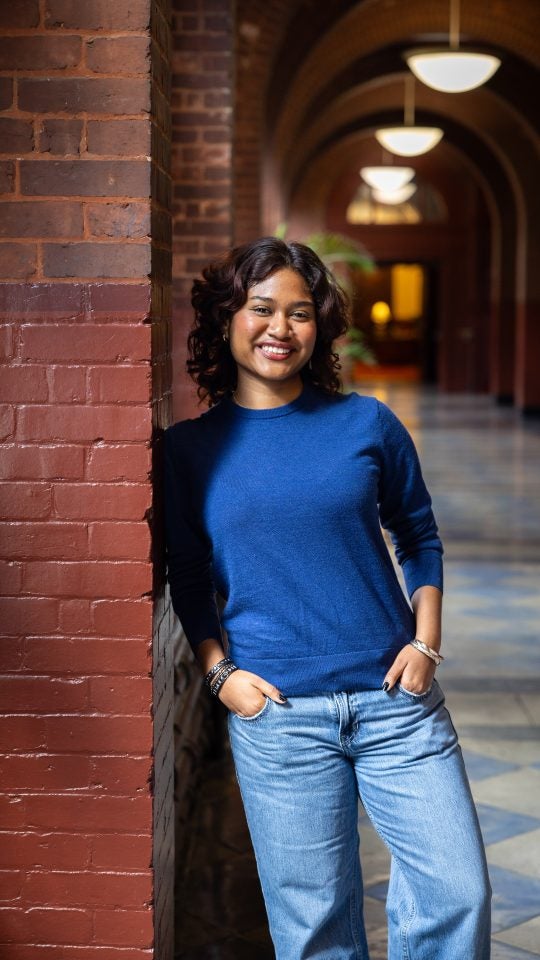 Young woman in a blue shirt leaning against a red brick pillar smiling with hands in jeans pocckets