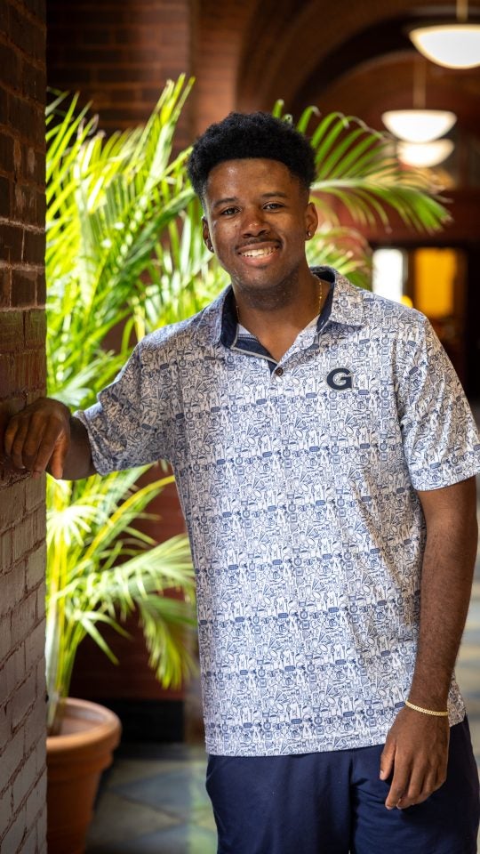 Young black man leans against a red brick pillar in a GU polo shirt smiling