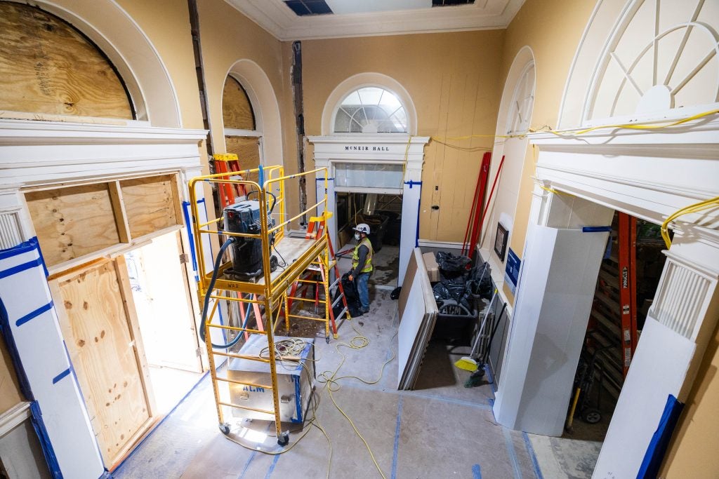 Construction on the inside of a building. A workman stands beside a ladder wearing a vest and hard hat.