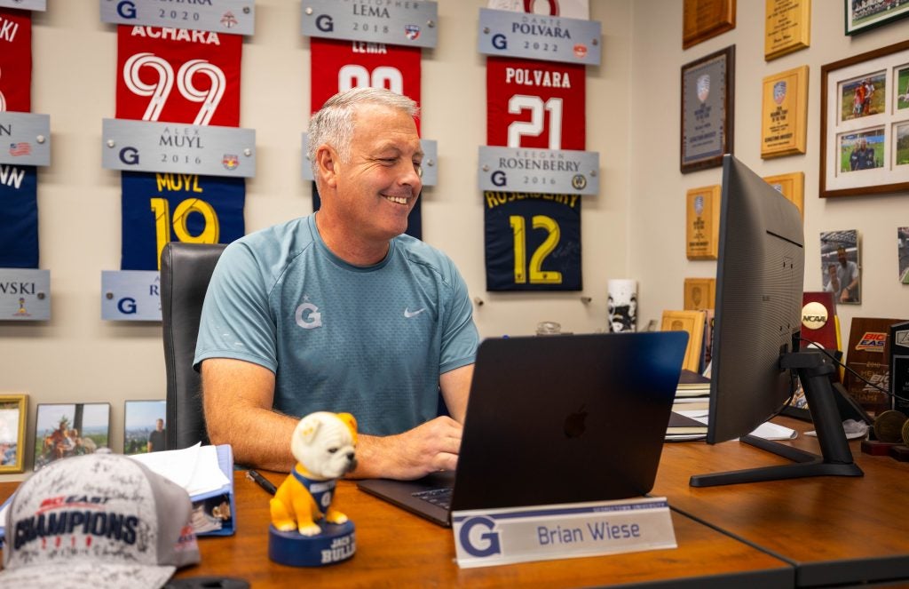 A coach smiles while working on his computer at a desk