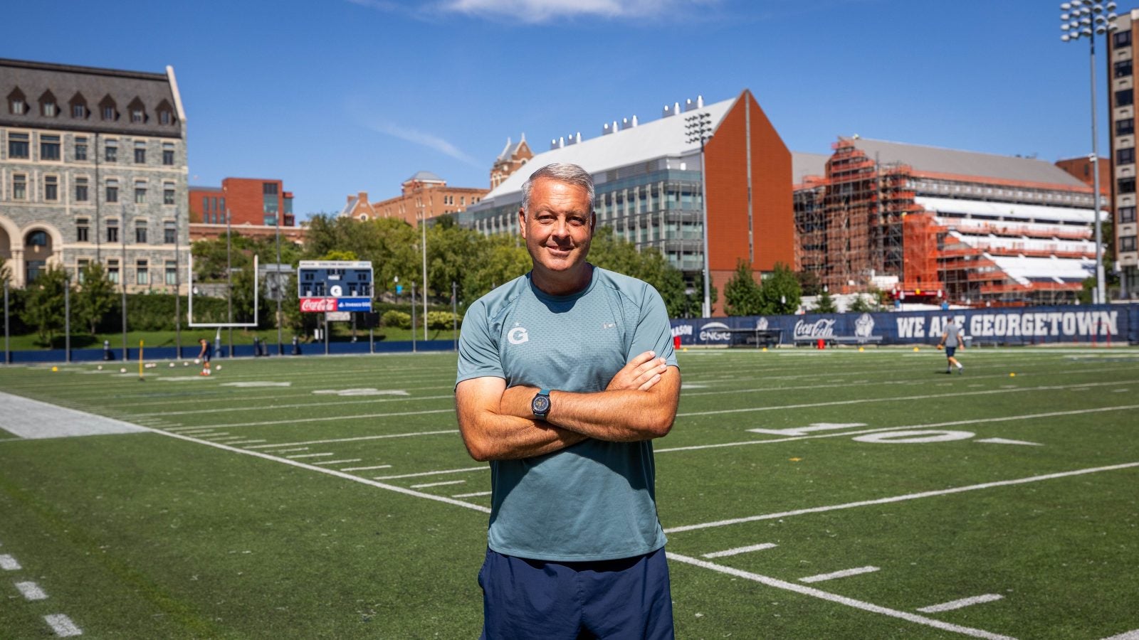 A coach in a blue GU shirt with arms crossed smiling while on field on a sunny day