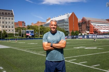 A coach in a blue GU shirt with arms crossed smiling while on field on a sunny day