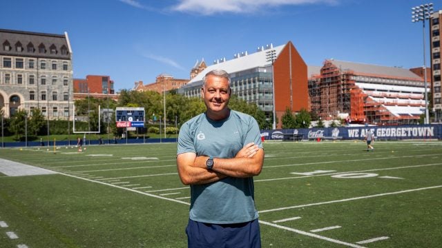 A coach in a blue GU shirt with arms crossed smiling while on field on a sunny day