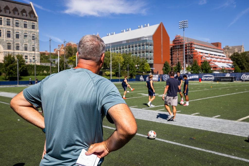 A coach watches his players warm up on a field