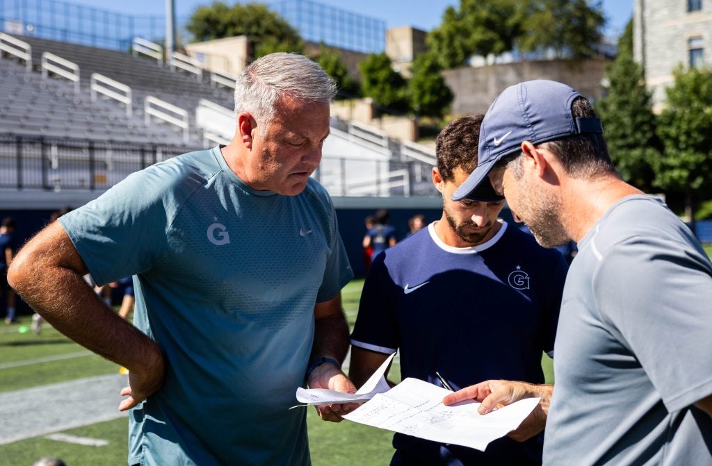 Three coaches look at a paper while standing on a field in the sun