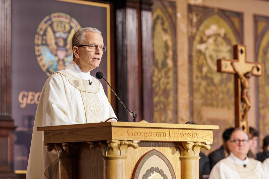 A Jesuit in a white robe stands in front of a podium next to a cross