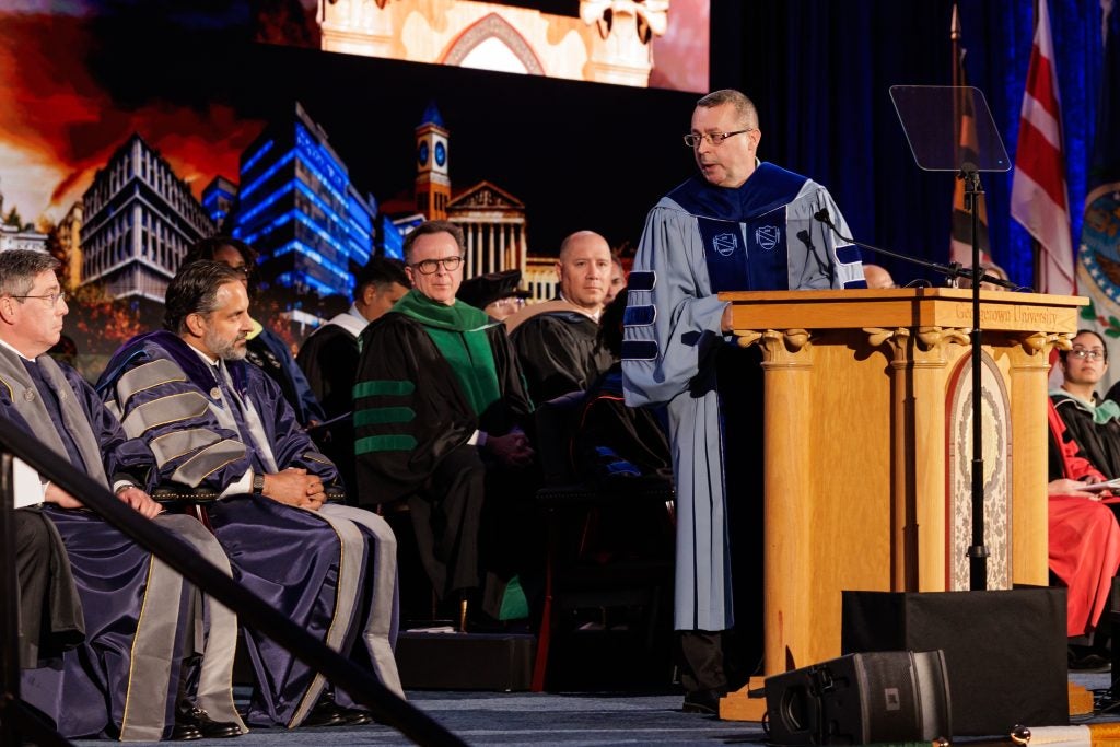 A professor in blue academic robes behind a podium speaks to Georgetown's president, who's seated onstage