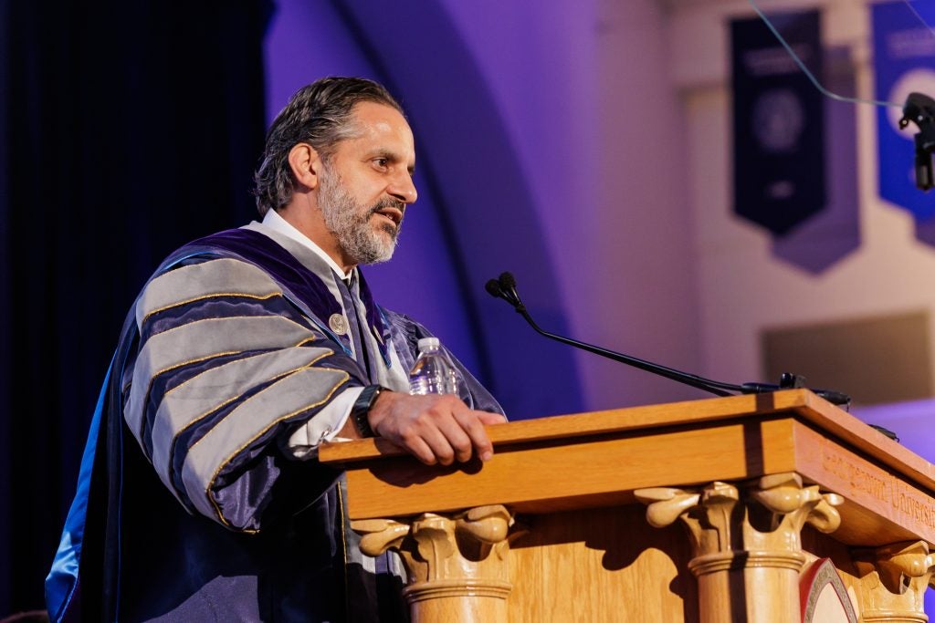 A man in a purple academic robe speaks from behind a podium