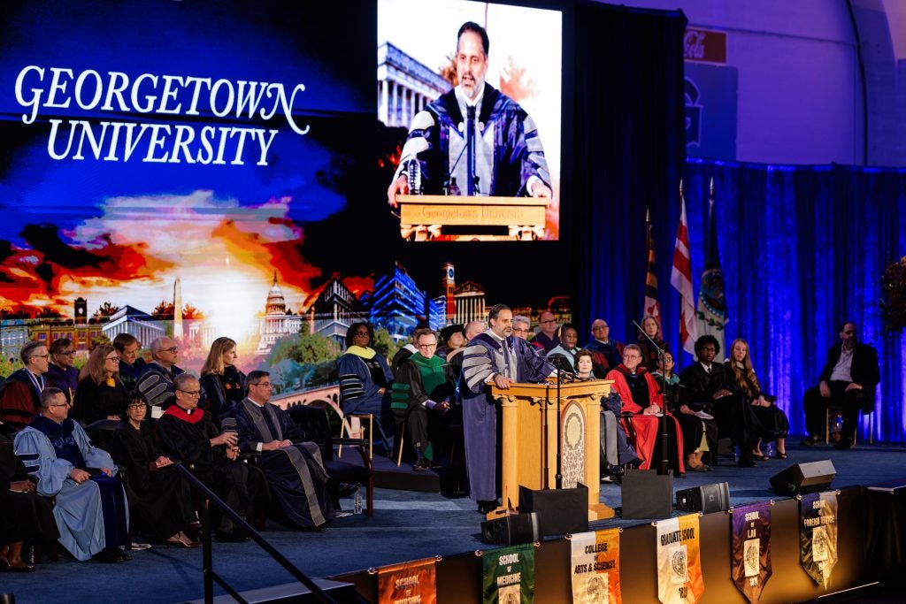 Georgetown's president speaks behind a podium with a live video feed projected above him