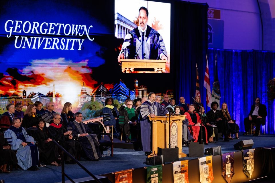 Georgetown's president speaks behind a podium with a live video feed projected above him