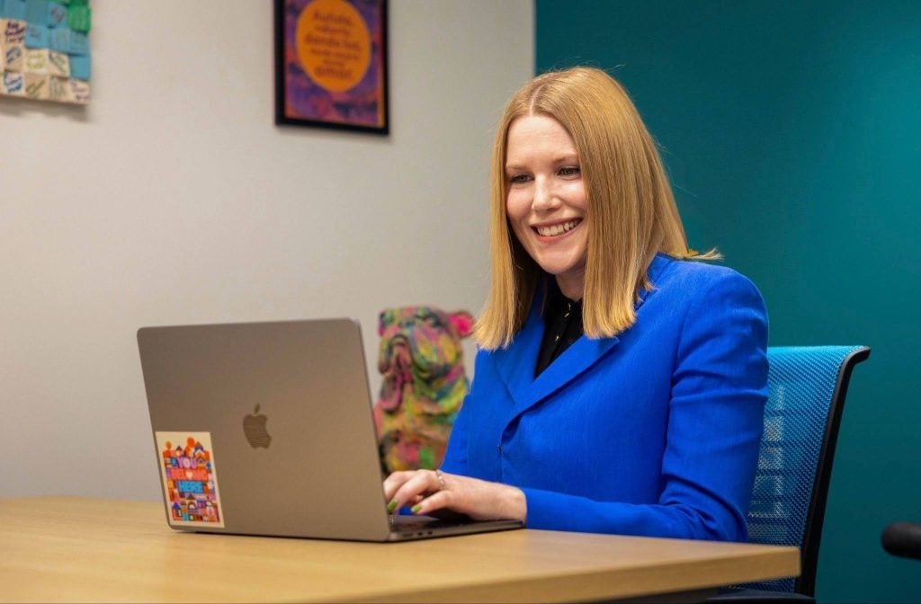 A woman in a blue blazer sits behind a desk and smiles while typing on her laptop