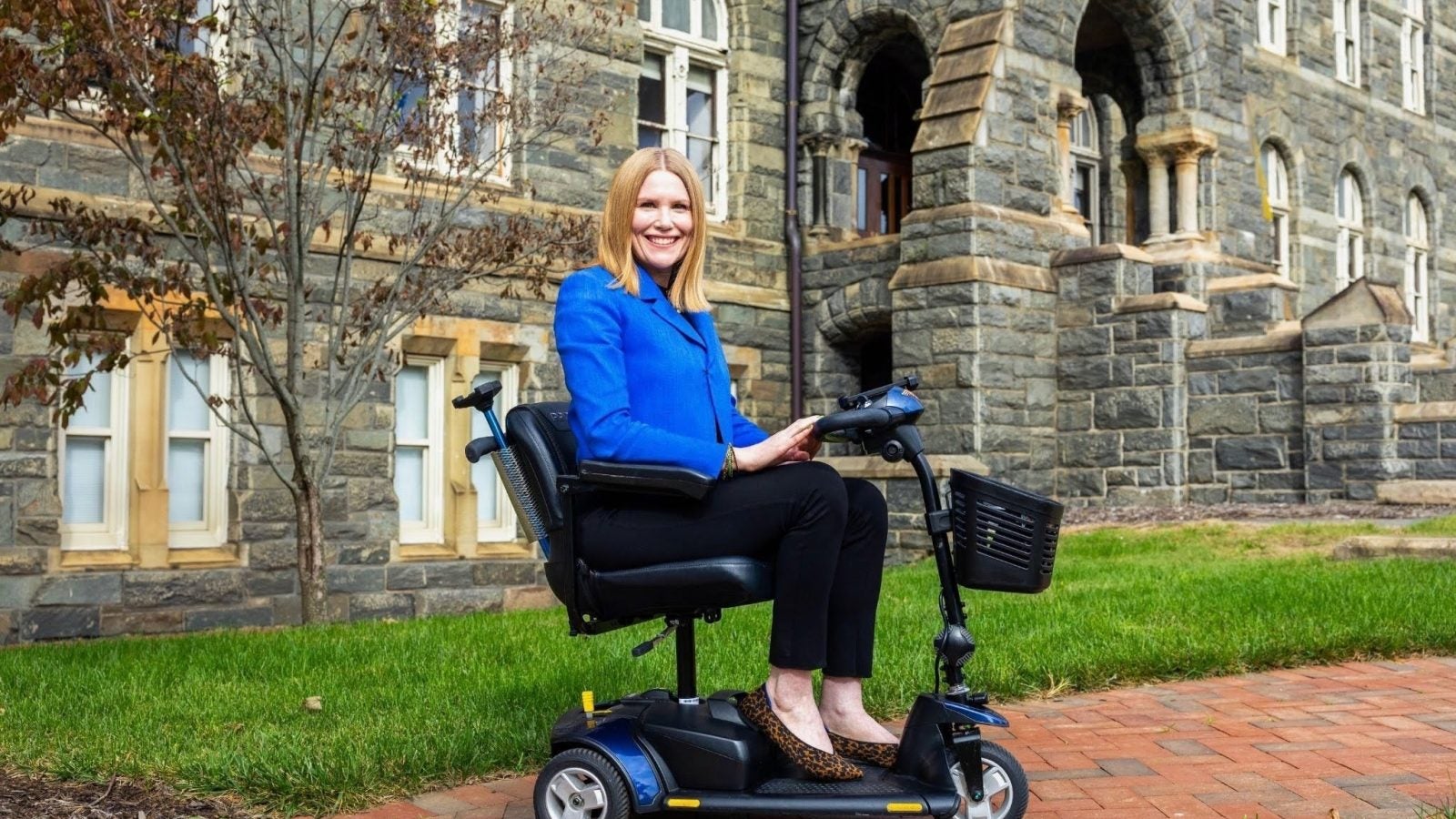 A woman in a blue blazer sits on a mobility scooter in front of a stone building