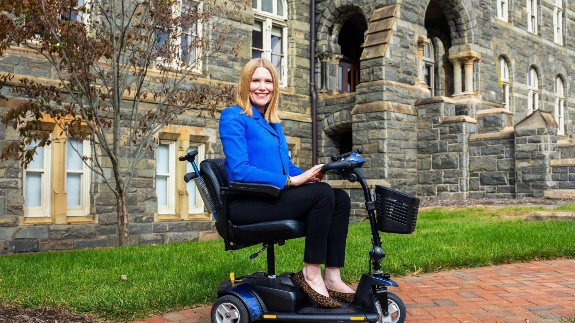 A woman in a blue blazer sits on a mobility scooter in front of a stone building