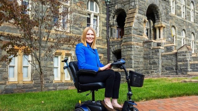 A woman in a blue blazer sits on a mobility scooter in front of a stone building