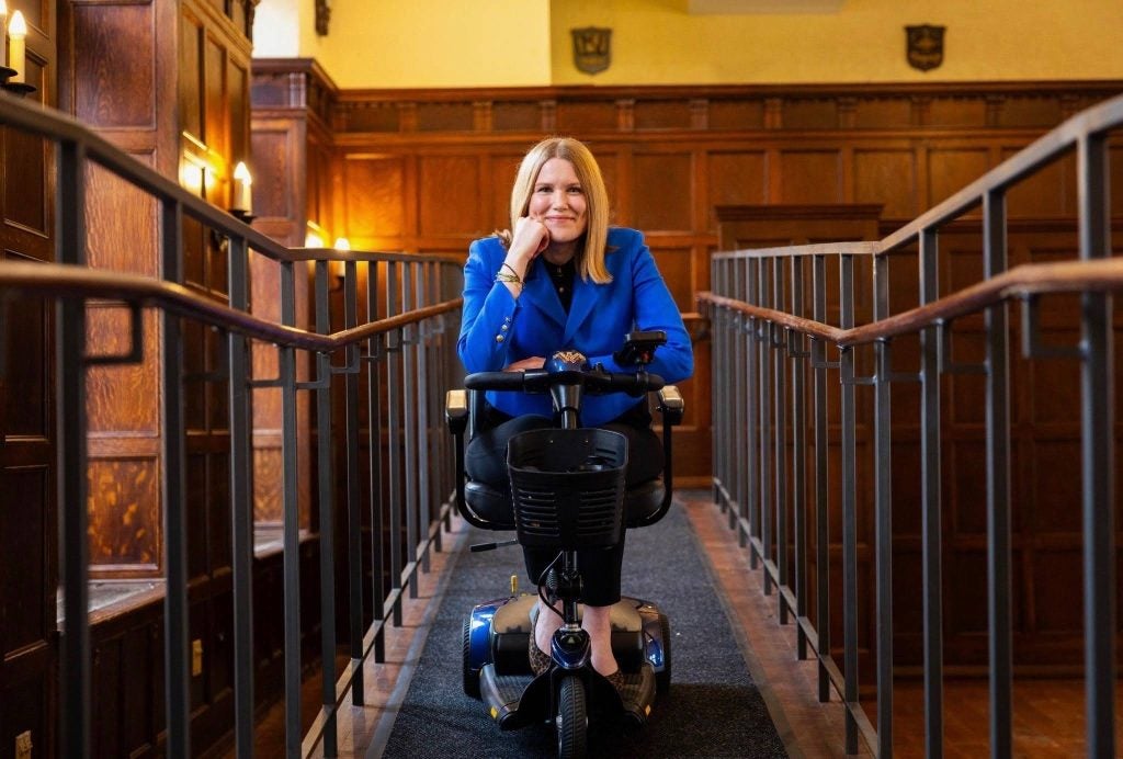 A woman on a mobility scooter rests her head on her hand on an indoor ramp. A wood paneled wall is behind her