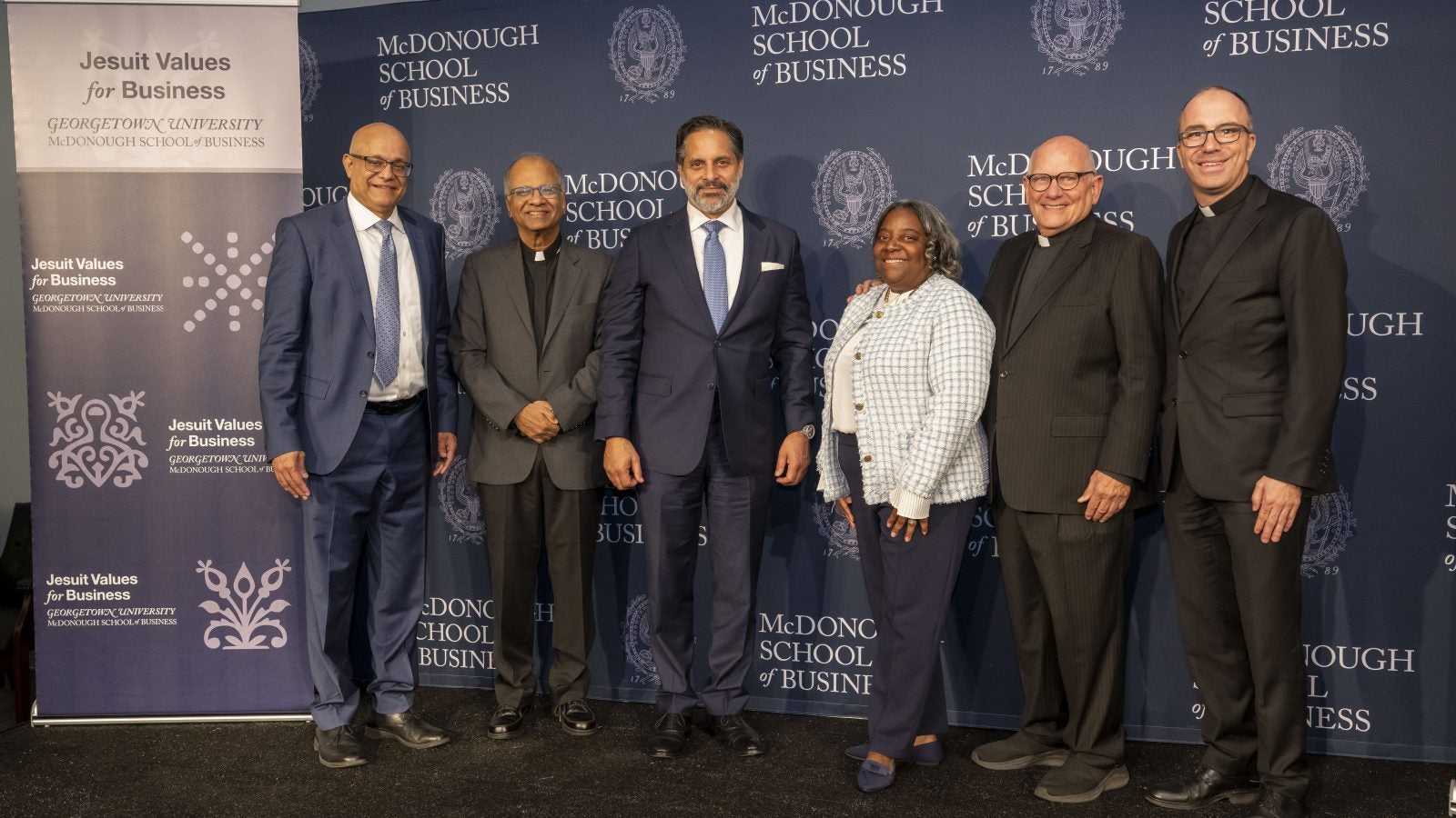 Jesuits, Business School leaders and Georgetown's president stand in front of a blue background.
