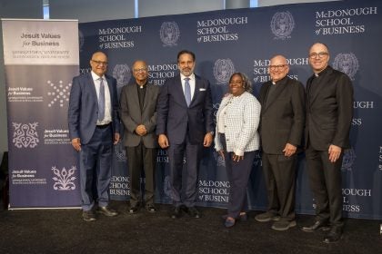 Jesuits, Business School leaders and Georgetown's president stand in front of a blue background.