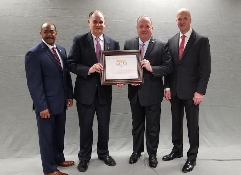 Four men stand in front of a gray background, with the two in the middle holding up the CALEA award.