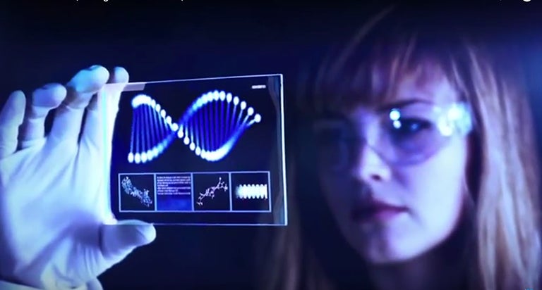 Woman in a white lab coat,goggles and gloves holds up a high-tech slide of a cell to the light as she conducts research 