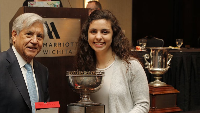 Greg Rosenbaum and Natalie Knez with trophy and other trophies and podium in the background 