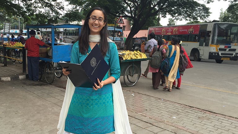 Chandini Jha (C'16), dressed in a bright turquoise dress, smiles and poses on a busy street in India. 