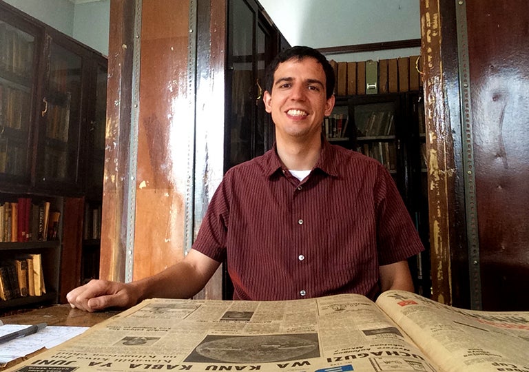 Andy Marshall sits at a desk with old periodicals on it and old wooden cases filled with books in the background.