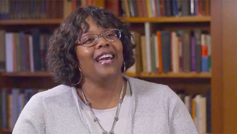 Kaya Henderson smiling wearing glasses with a library of books behind her stacked on shelves