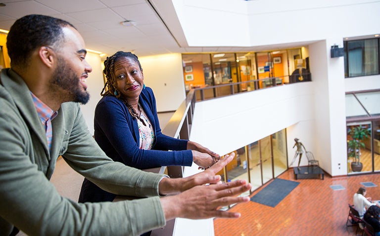 Professor Rosemary Ndubuizu looks at Professor Marcus Board as they lean over a railing chatting inside a camnpus building