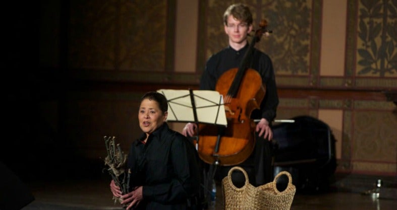 Anna Deavere Smith sitting on stage in Gaston Hall with Joshua Roman behind her holding a cello